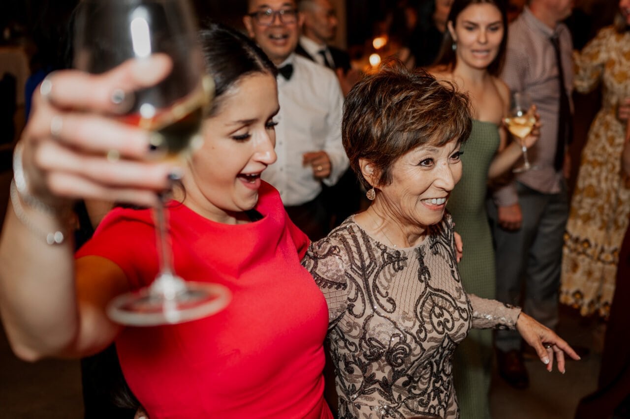 Two women, beaming with joy, dance at a lively party, drinks in hand. In the background, the festive atmosphere of an Amaterra Winery wedding reception unfolds.