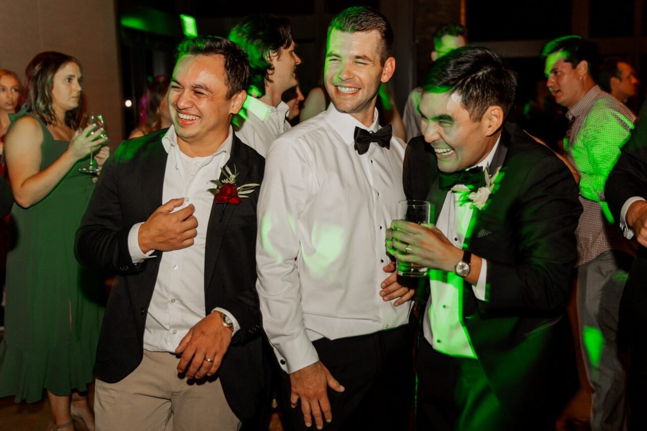 Three men in formal wear are laughing at a lively party, capturing the joyous essence of an Amaterra Winery wedding. In the background, guests dance and mingle, adding to the festive atmosphere of this memorable occasion.