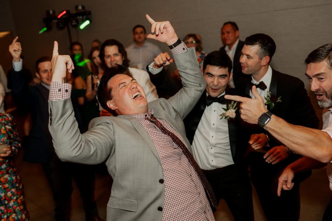 A joyful person in a suit is dancing enthusiastically at a lively celebration, surrounded by smiling people capturing the blissful moments, as if posing for Amaterra Winery wedding photos.