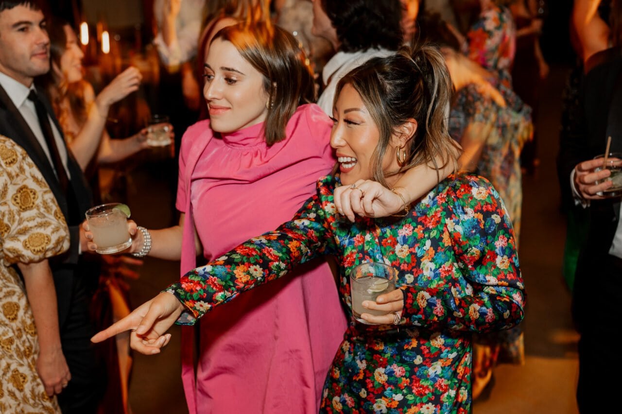Two women in colorful dresses dance and laugh, holding drinks, as the vibrant atmosphere of an Amaterra Winery wedding swirls around them. Other guests are delightfully blurred in the background, capturing a moment of pure joy and celebration.