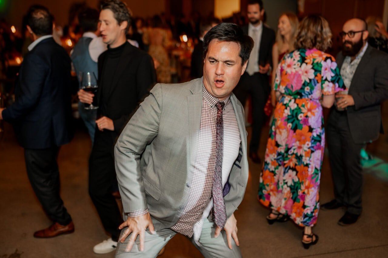 A man in a gray suit dancing energetically at a lively party, perhaps captured in Amaterra Winery wedding photos, with people joyfully socializing in the background.