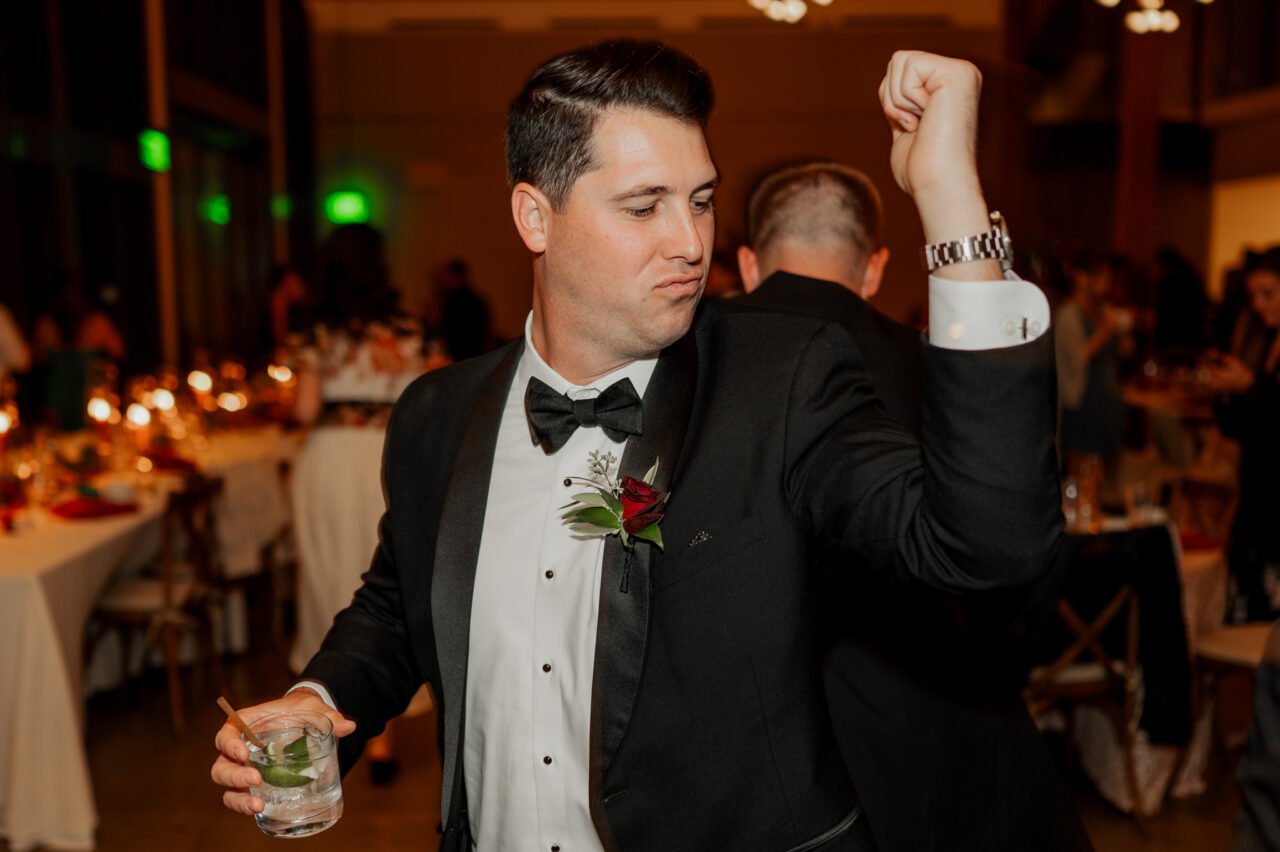 In the dimly lit banquet hall, a man in a suit flexes his arm while holding a drink, capturing the sophisticated charm reminiscent of Amaterra Winery wedding photos.