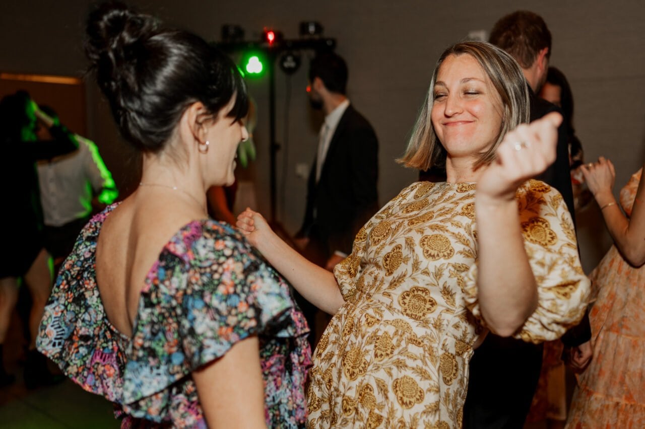 Two women smiling and dancing at a party set against the elegant backdrop of Amaterra Winery, surrounded by guests in semi-formal attire, capturing the joy found in wedding photos.