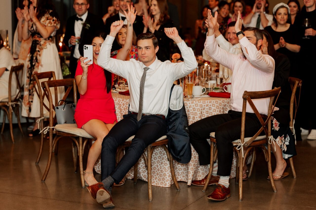 Guests clapping and cheering at an indoor event, reminiscent of joyful moments captured in Amaterra Winery wedding photos, with a man seated in the foreground holding his hands up.