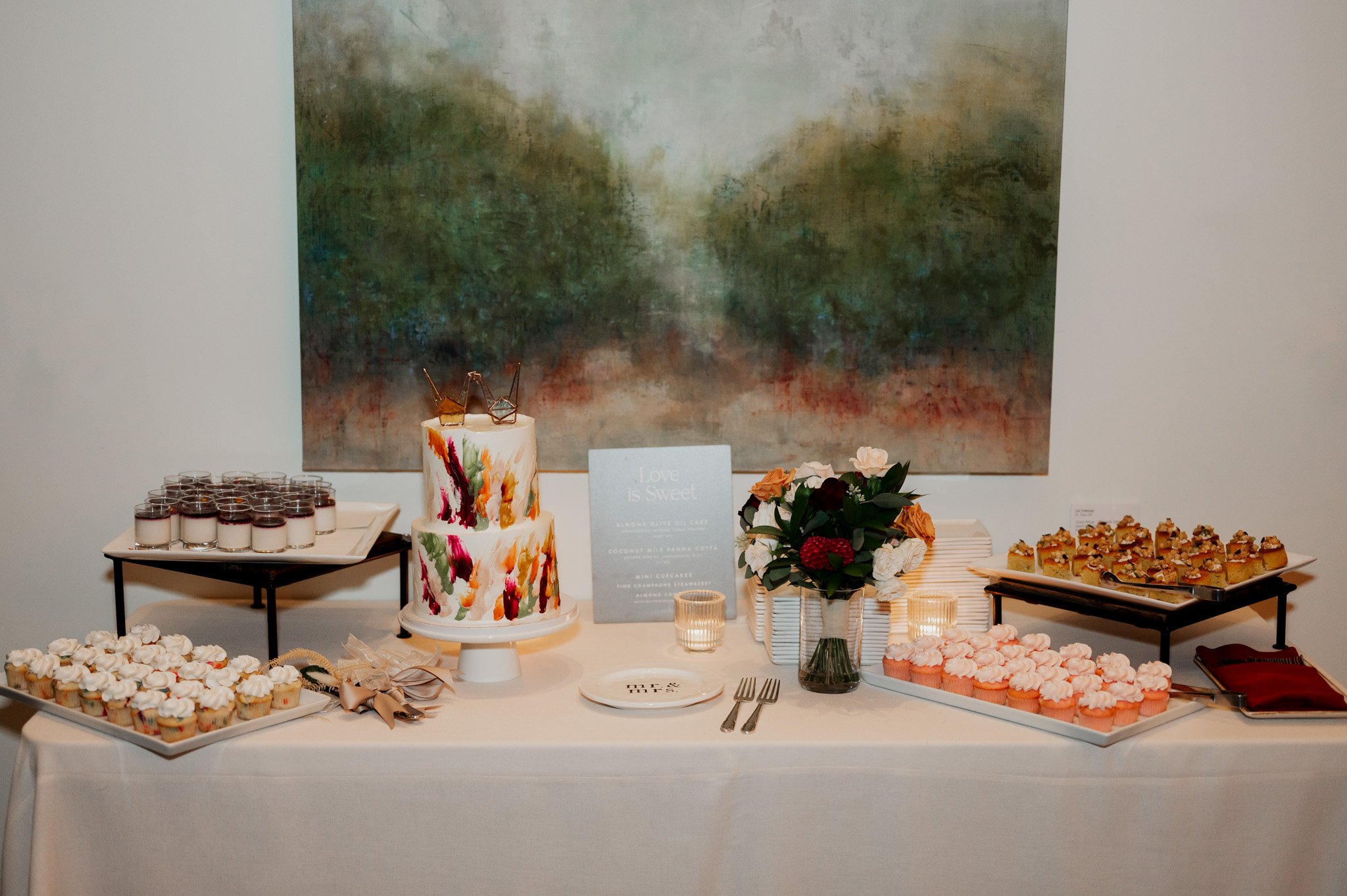A dessert table featuring a colorful cake, cupcakes, and pastries is beautifully set against a landscape painting backdrop. With flowers and a decorative sign, its perfect for capturing Amaterra Winery wedding photos.
