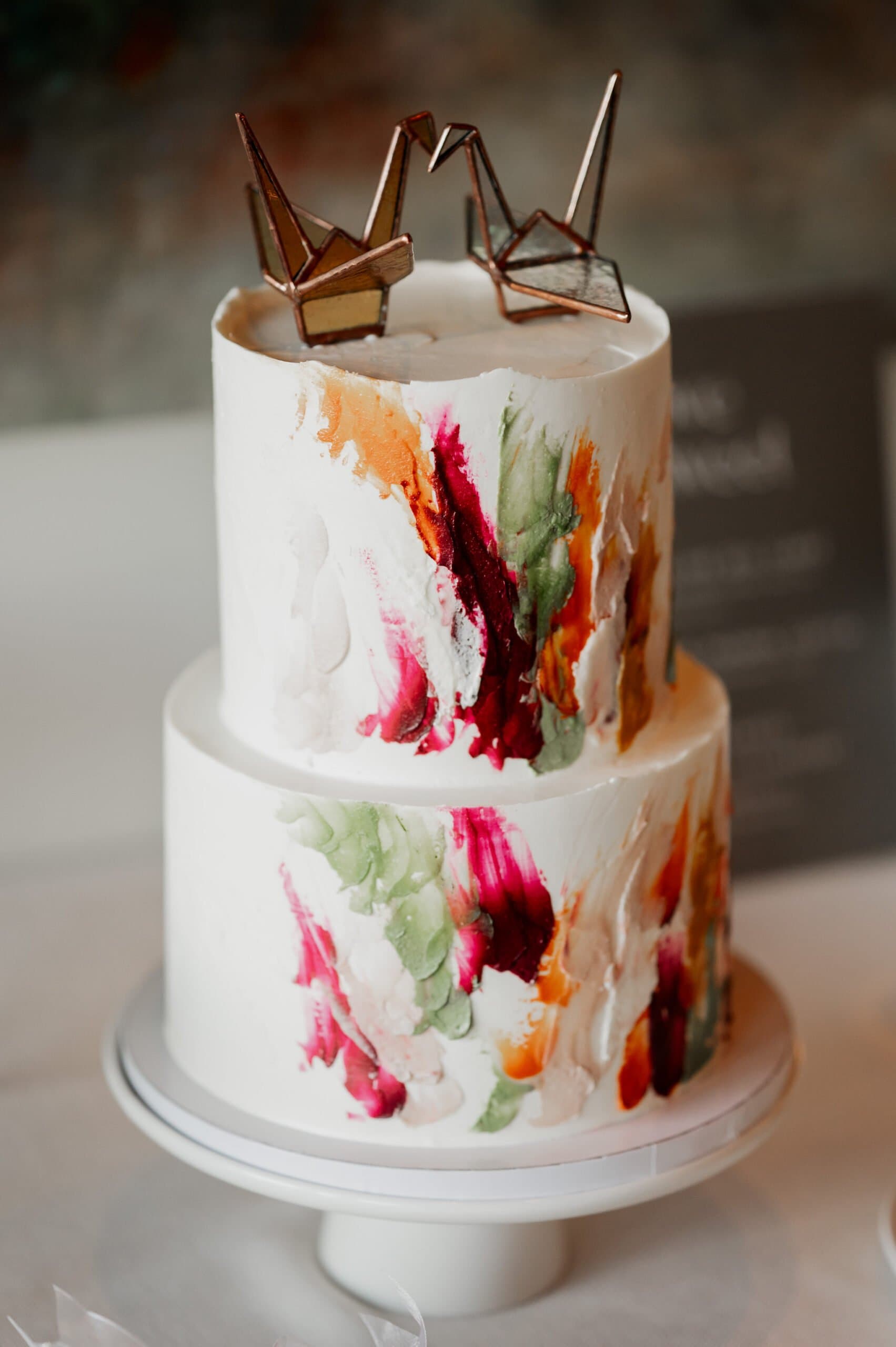 A two-tier white cake adorned with colorful brushstroke designs and topped with two origami cranes, perfect for capturing in Amaterra Winery wedding photos.