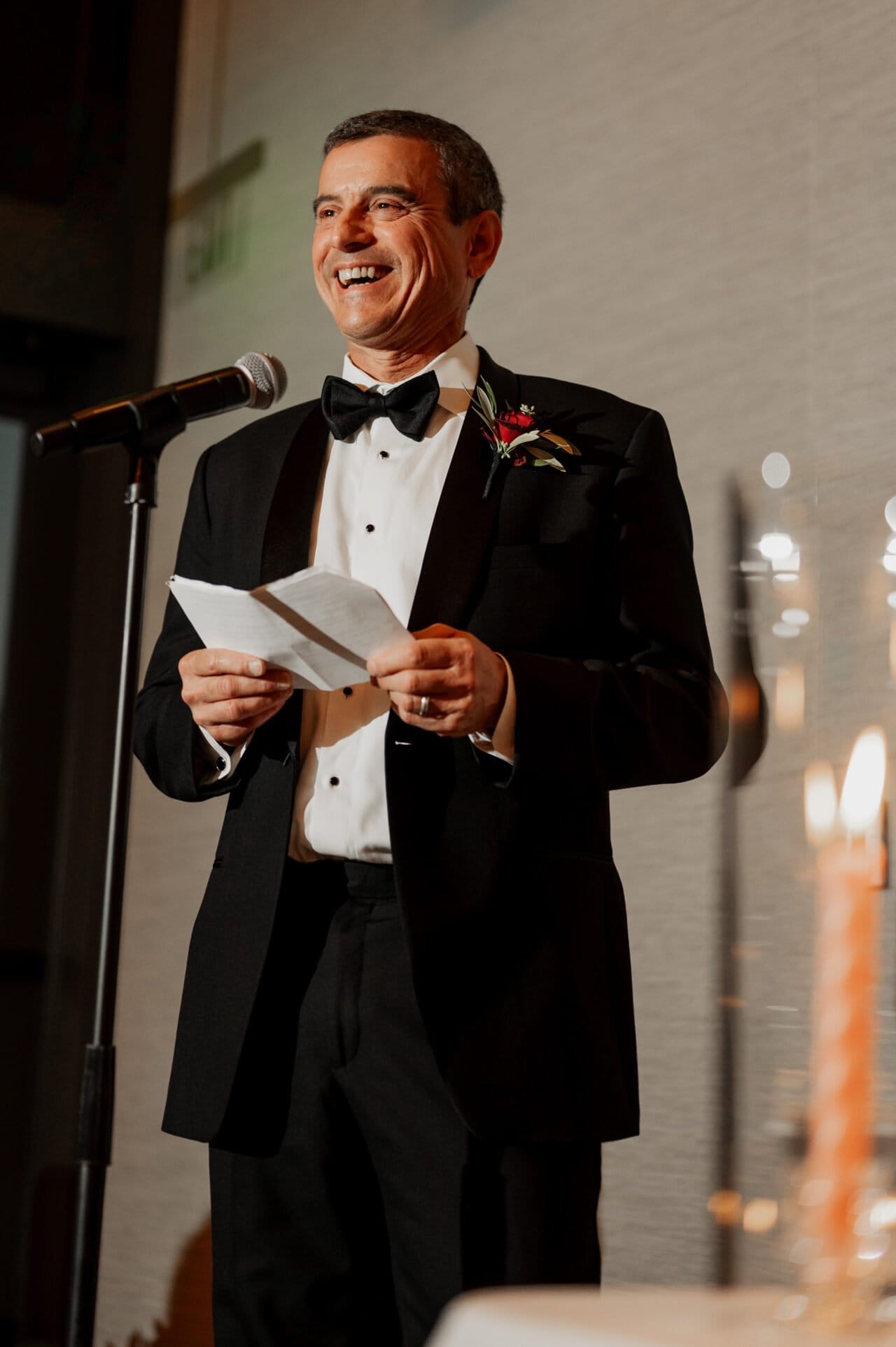 Man in tuxedo, smiling as he speaks into a microphone at an Amaterra Winery wedding. A piece of paper in hand and nearby candles bathe the moment in a romantic glow.