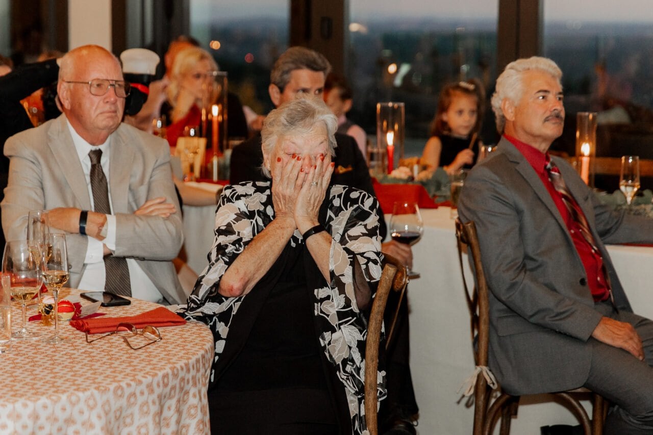 An elderly woman covers her face while seated at a formal event, surrounded by other attendees, capturing a poignant moment at the Amaterra Winery wedding.