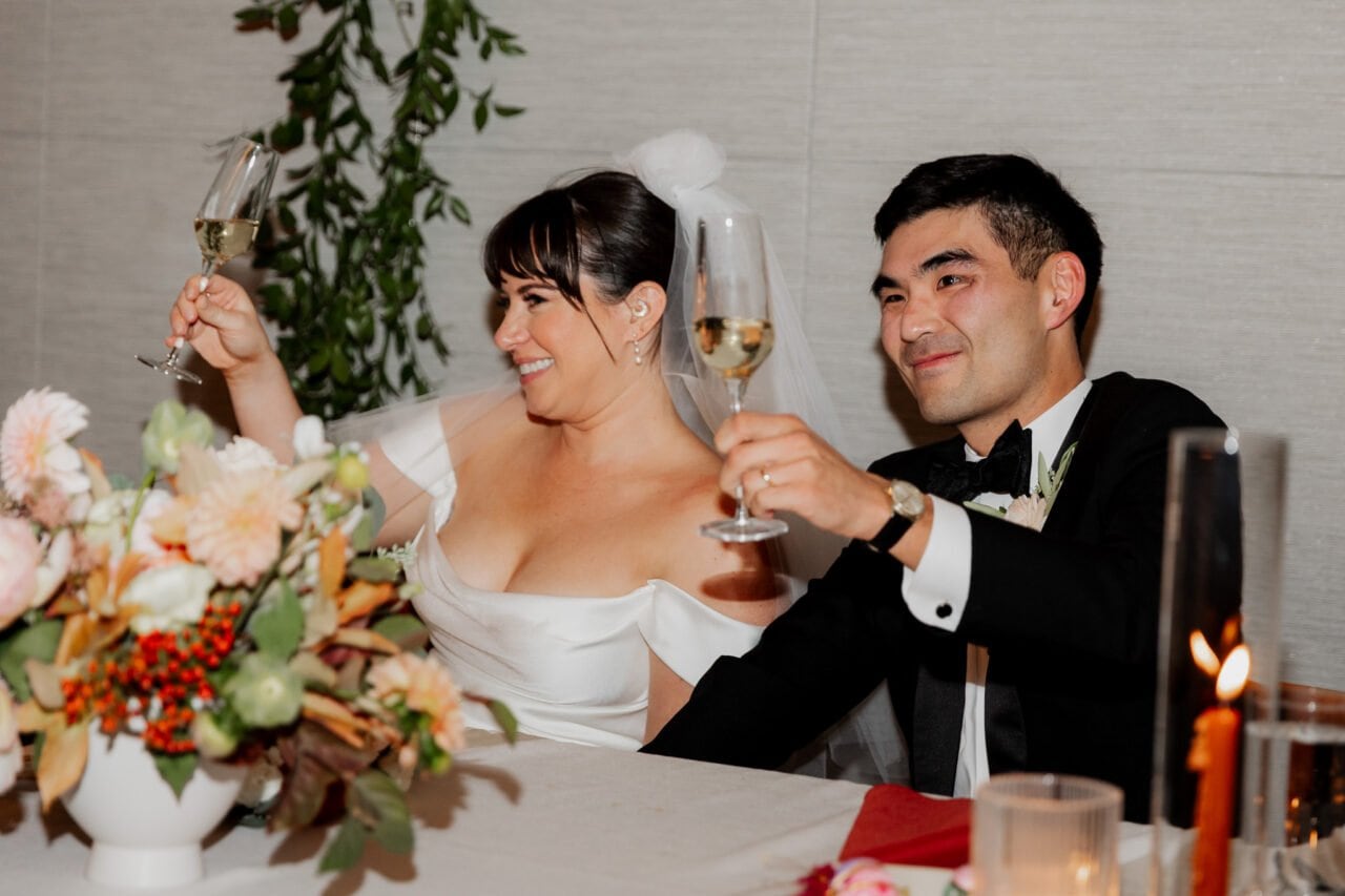 A couple in wedding attire toasts with champagne, seated at a beautifully decorated table amidst the stunning backdrop of Amaterra Winery, smiling joyfully.