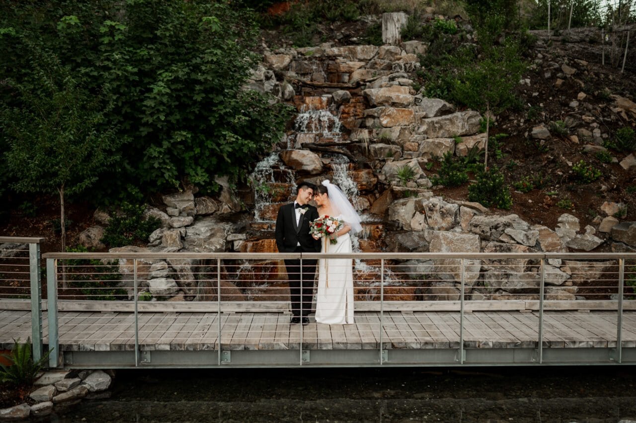 Bride and groom embrace on a bridge in front of a rocky waterfall and lush greenery, capturing the enchanting essence of Amaterra Winery wedding photos.