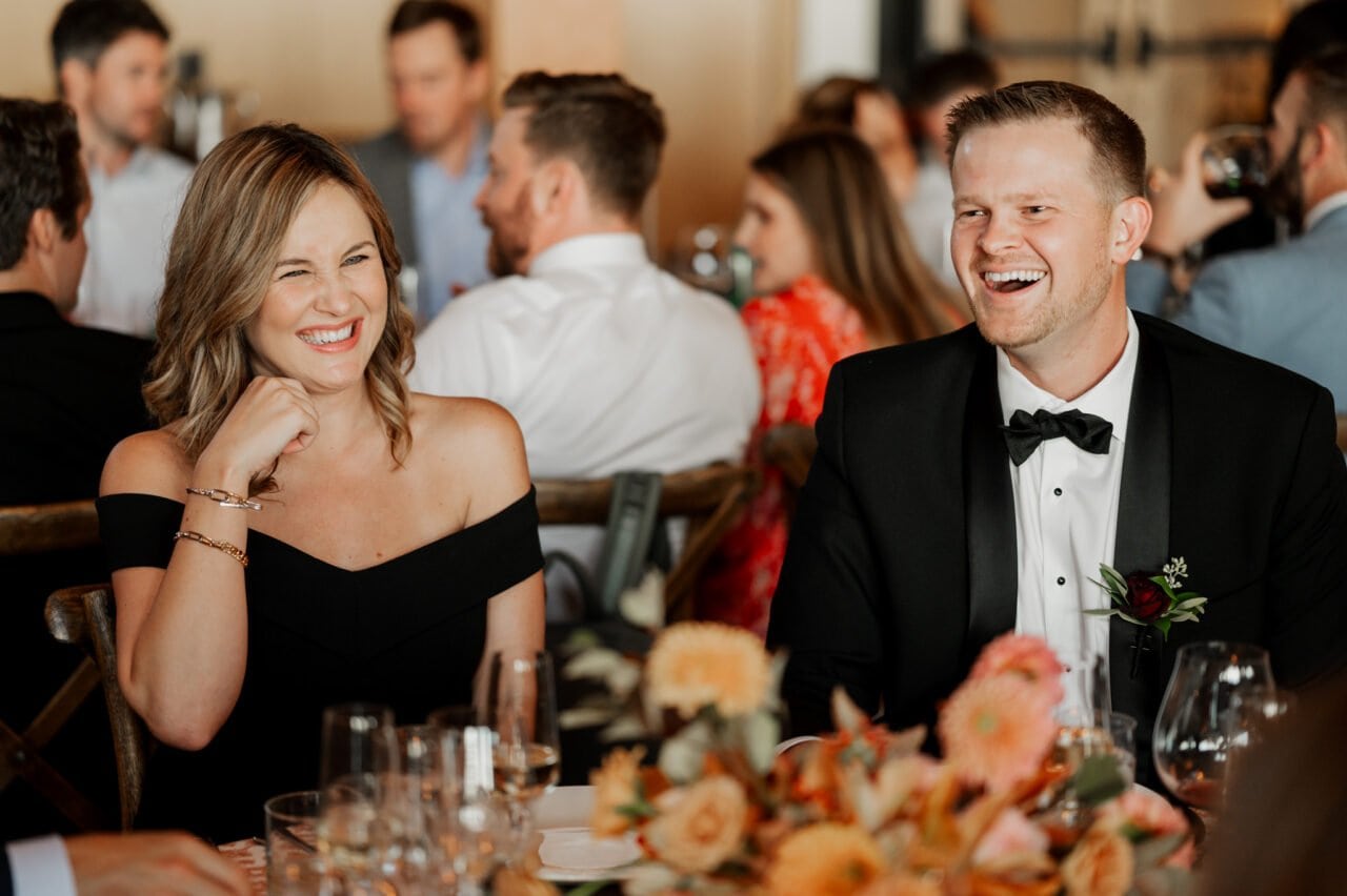 A smiling woman in a black dress and a man in a tuxedo enjoy a formal event at Amaterra Winery, with others seated at tables, capturing the essence of an enchanting wedding celebration.
