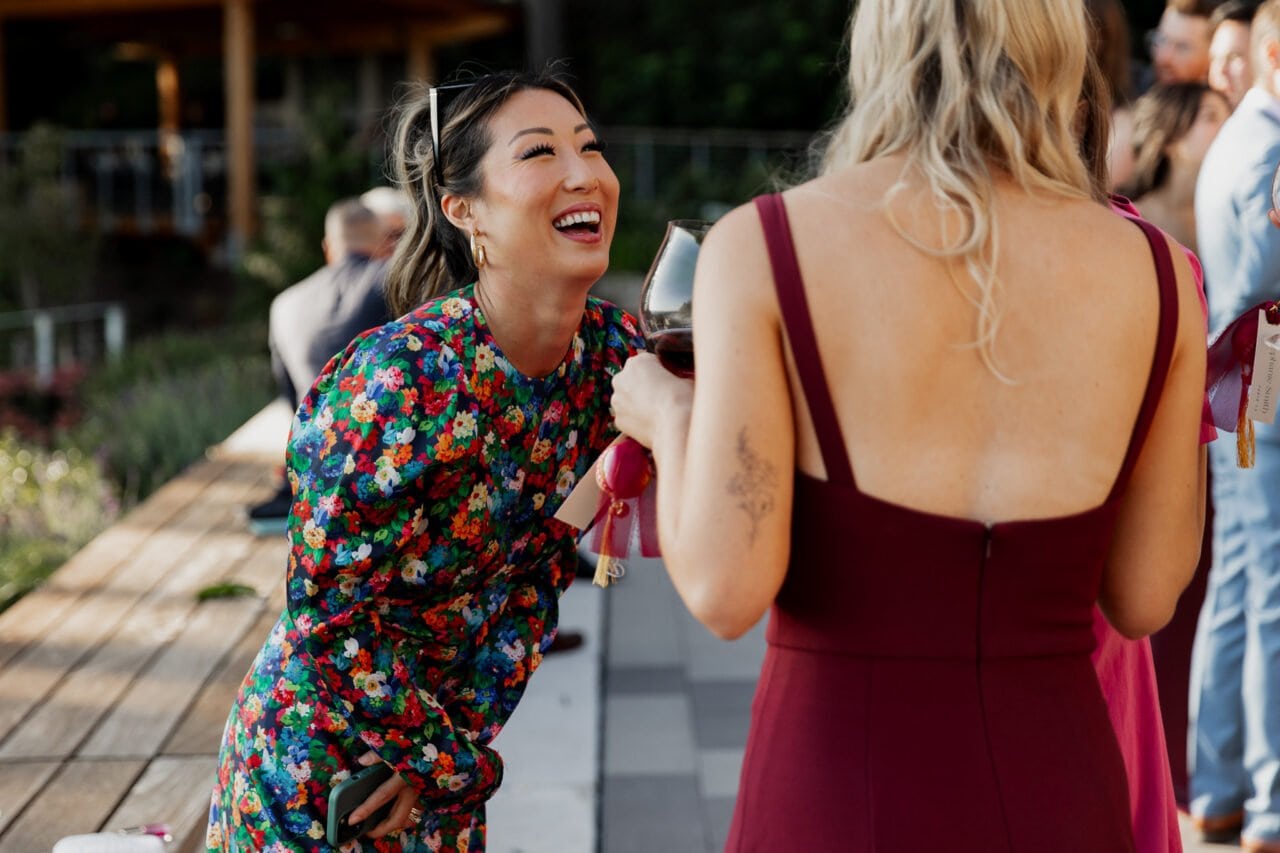 Two people laughing, one in a floral dress and the other in a burgundy dress, each holding a wine glass under the sun at an Amaterra Winery wedding.