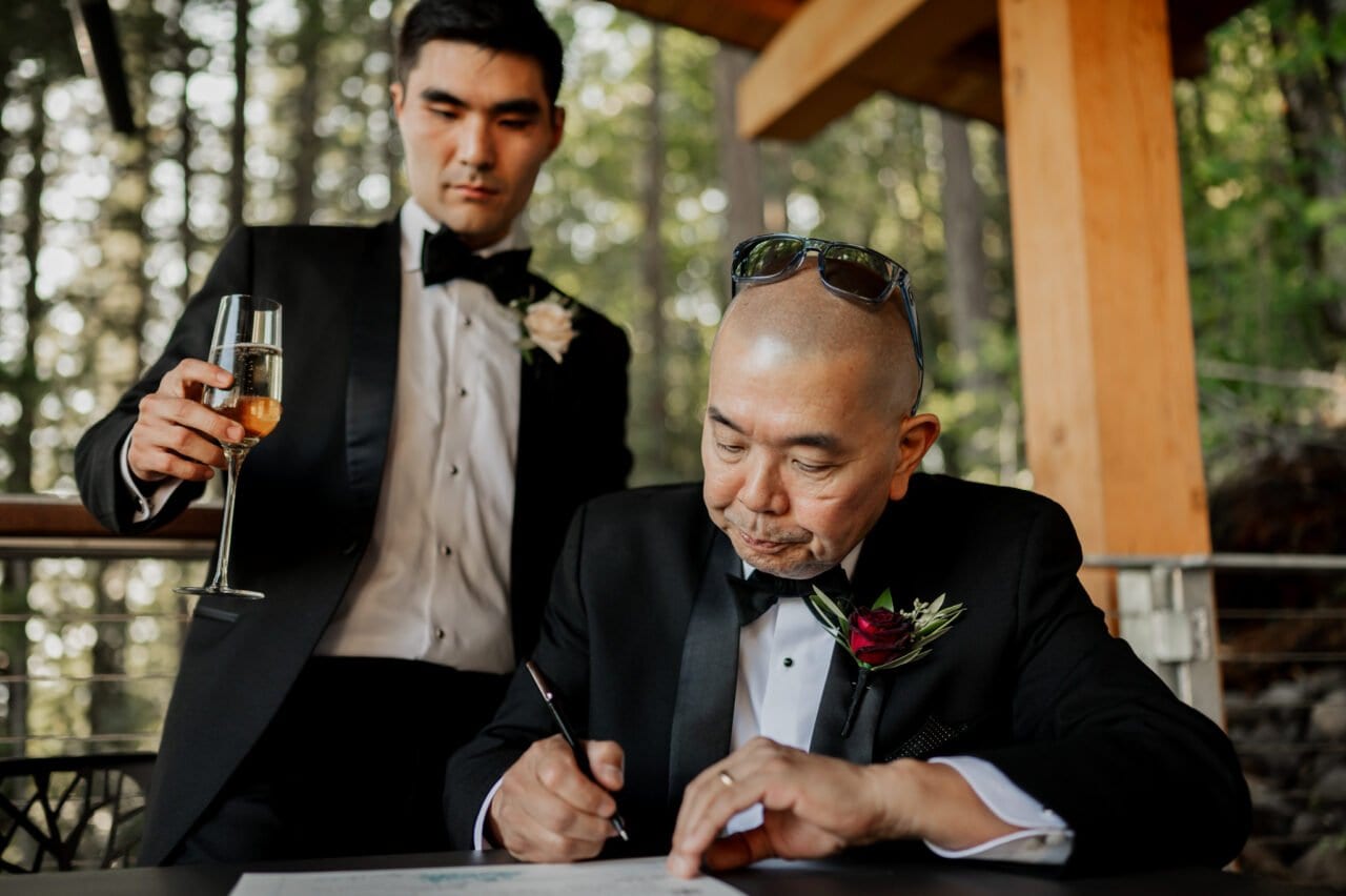 In a charming forest setting reminiscent of Amaterra Winery wedding photos, two men in tuxedos create a perfect moment. One signs a document while the other elegantly holds a champagne glass, capturing the essence of celebration and commitment amidst natures beauty.