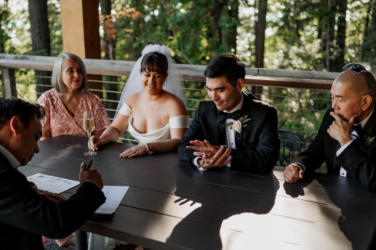 Amidst the picturesque backdrop of Amaterra Winery, a bride and groom happily sign their wedding document, surrounded by joyful family members in an enchanting outdoor setting.