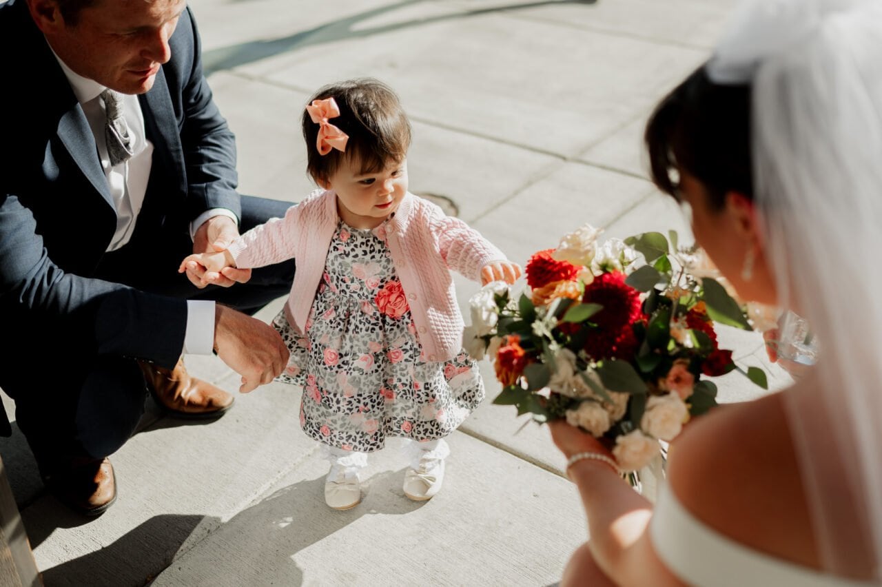 During an Amaterra Winery wedding, a bride gracefully shows flowers to a toddler in a floral dress, lovingly held by a man in a suit under the open sky.