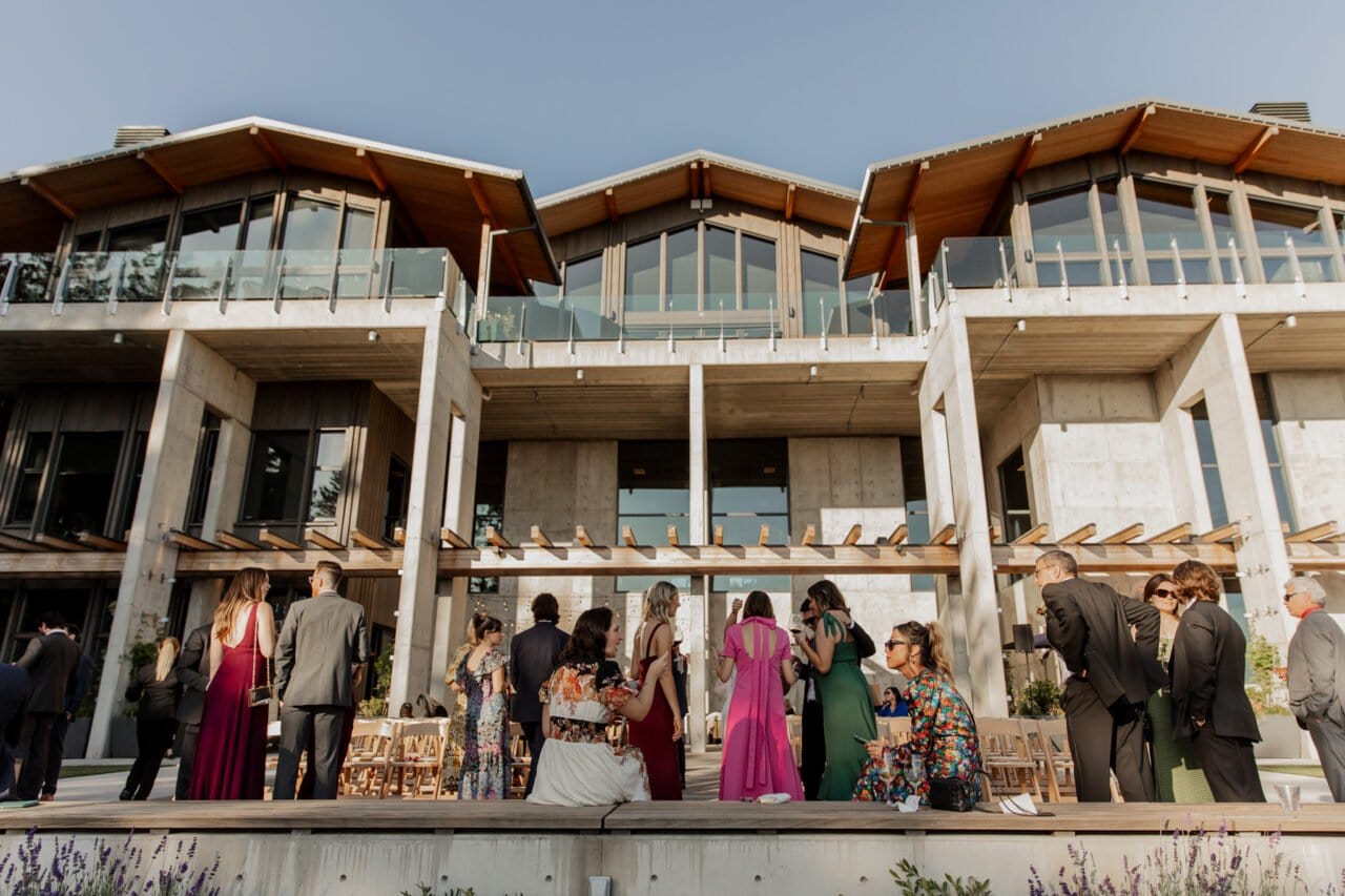 Guests in formal attire gather outside a modern building with large glass windows and wooden roofs, capturing perfect Amaterra Winery wedding photos.