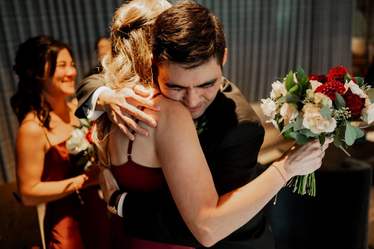 A man in a suit hugs a woman holding a bouquet at the Amaterra Winery. Another woman in a red dress smiles in the background, capturing the joy of wedding photos at this enchanting venue.
