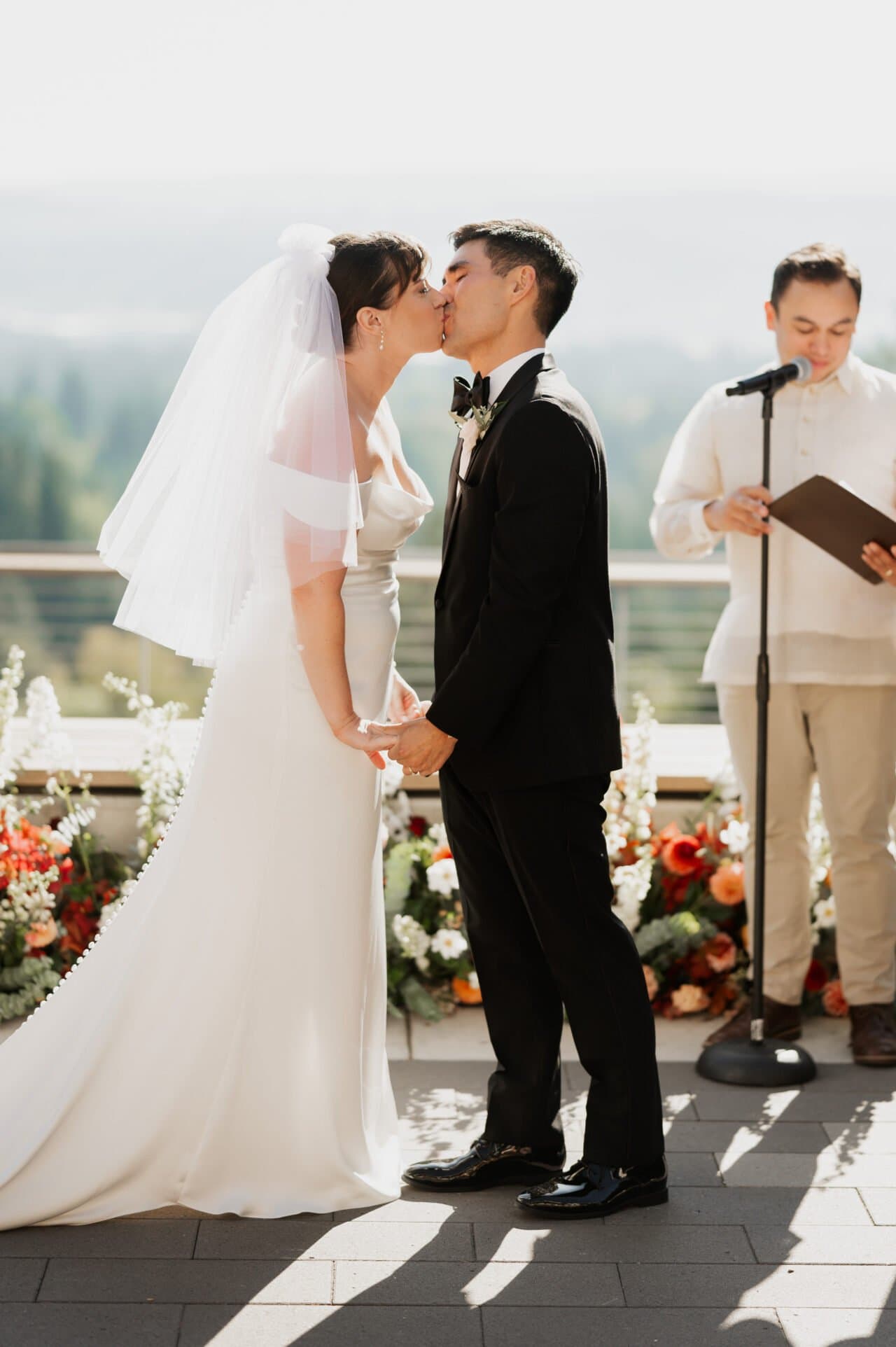 During their Amaterra Winery wedding, a bride and groom share a kiss amidst the lush vines, as the officiant stands close by, capturing a timeless moment.