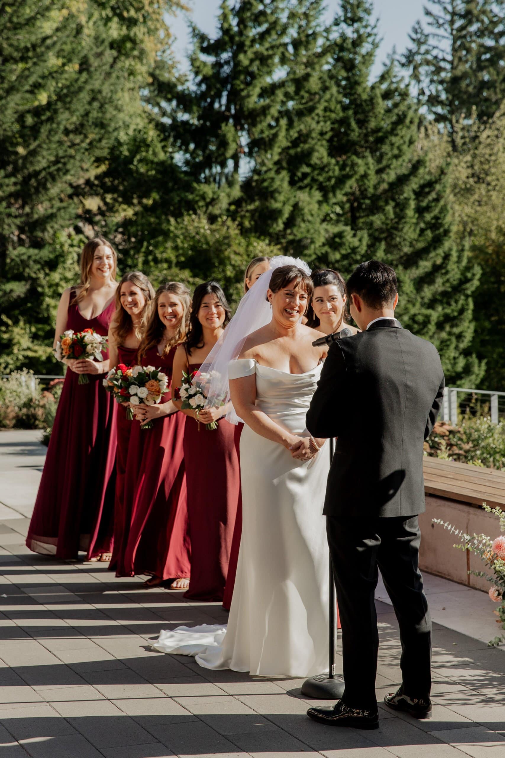 At the beautiful outdoor wedding ceremony, the bride and groom stand with bridesmaids in burgundy dresses by their side, enveloped by trees. The scene captures the magic of Amaterra Winery wedding photos.