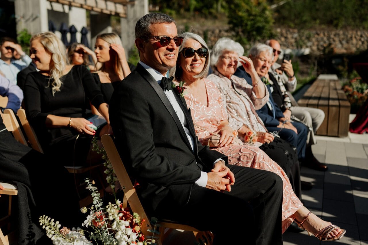 At Amaterra Winery, people in formal attire sit outdoors, smiling. A man in a suit and a woman in a pink dress are captured beautifully in the foreground, embodying the joy of winery wedding photos.