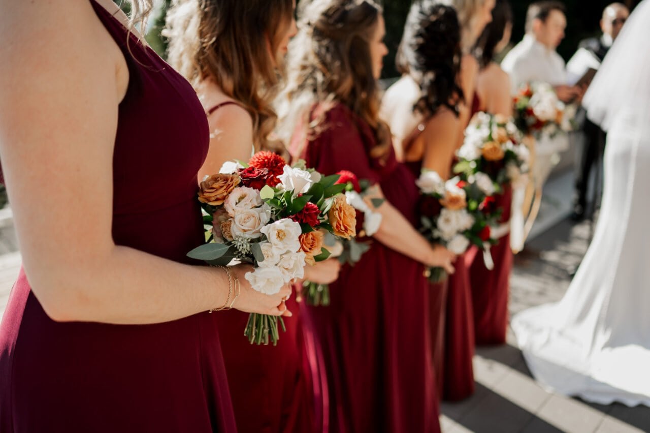 Bridesmaids in burgundy dresses hold bouquets, their vibrant colors beautifully captured in Amaterra Winery wedding photos during an enchanting outdoor ceremony.
