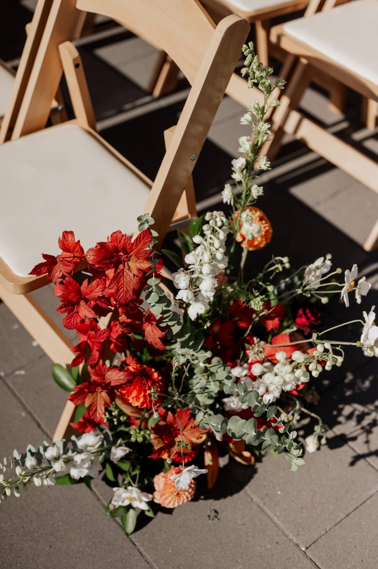 A wooden chair, adorned with a vibrant bouquet of red, white, and orange flowers, graces the sunny stone pathway—a scene reminiscent of enchanting Amaterra Winery wedding photos.