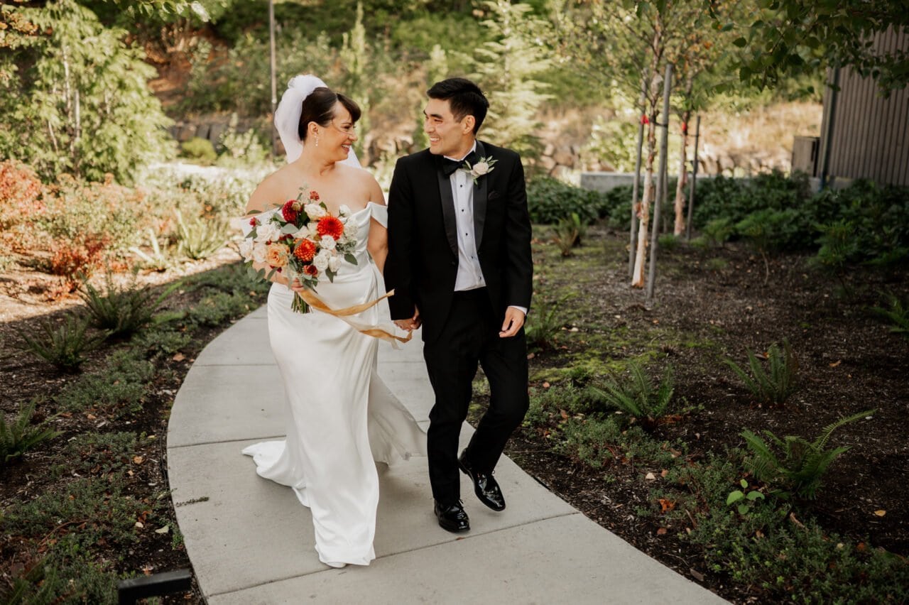 The bride and groom joyfully walk hand in hand on a garden path at Amaterra Winery, surrounded by lush greenery and the beauty captured in their wedding photos.