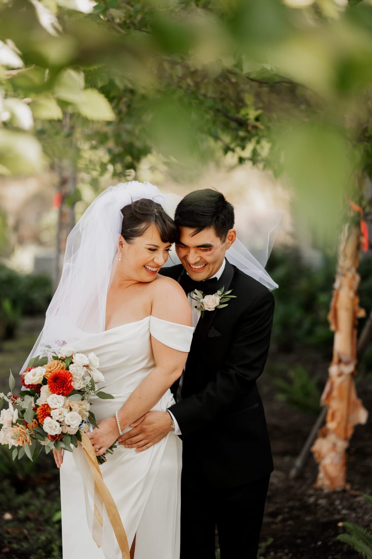 Bride and groom smiling under leafy trees at Amaterra Winery, bride holding a colorful bouquet.