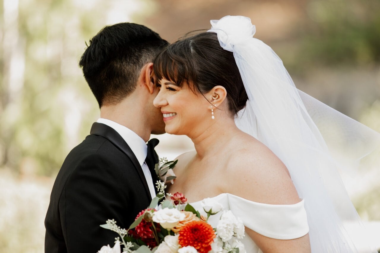 Bride in white gown smiling, holding bouquet, standing with groom in black suit at Amaterra Winery; a perfect setting for wedding photos under the sunny sky.