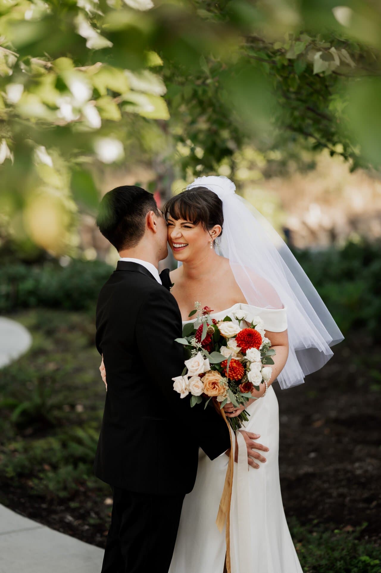 A bride in a white dress and veil beams as she holds red and white flowers, embracing her groom in a black suit at the picturesque Amaterra Winery.