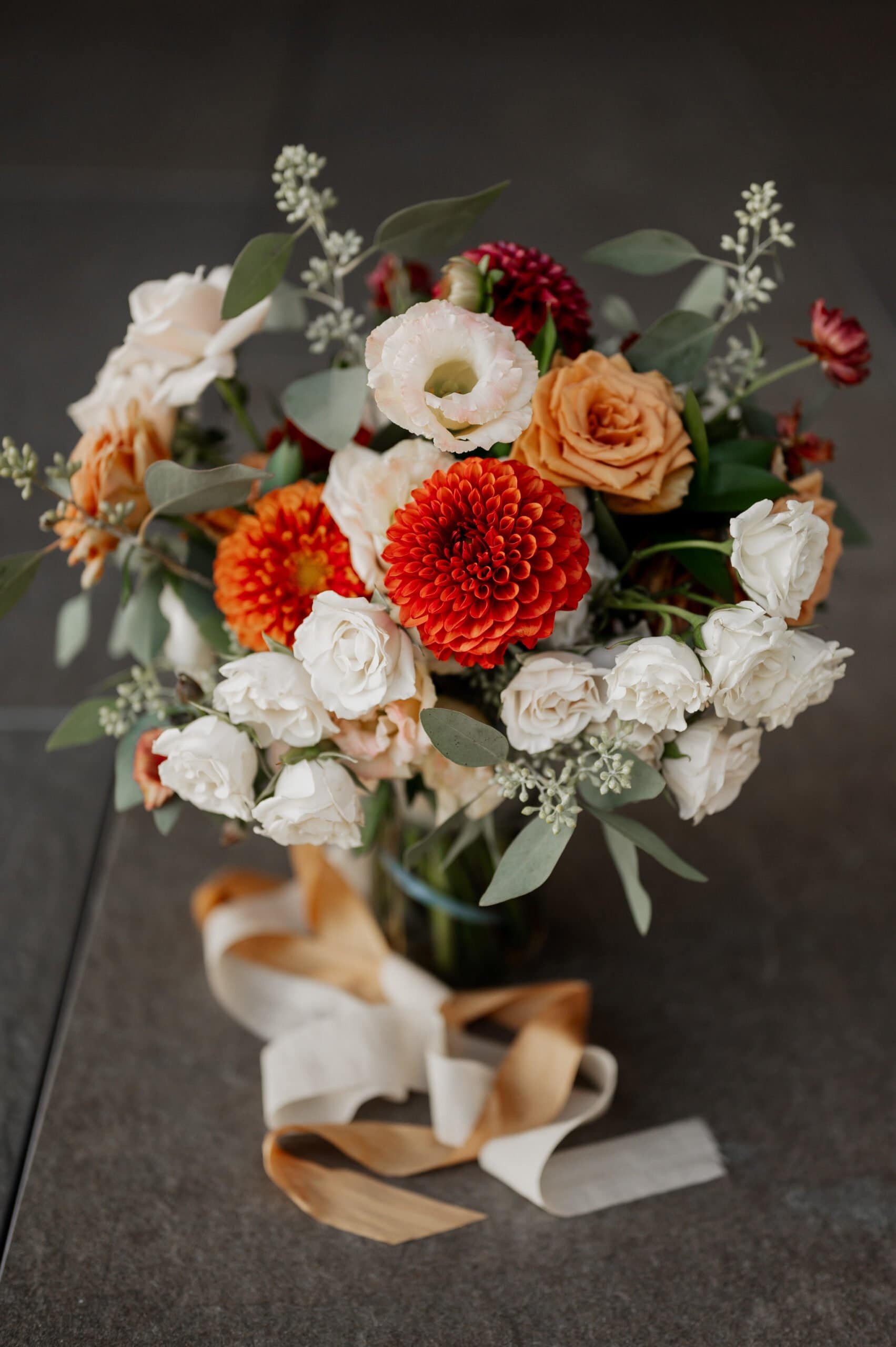 A bouquet of white, orange, and blush flowers with green leaves and ribbon rests elegantly on a dark surface, reminiscent of Amaterra Winery wedding photos.