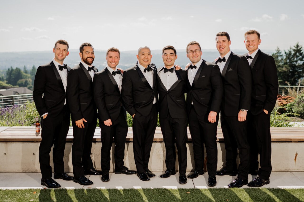 Eight men in tuxedos smile outdoors against the scenic backdrop of Amaterra Winery, capturing the joy and elegance of wedding photos.