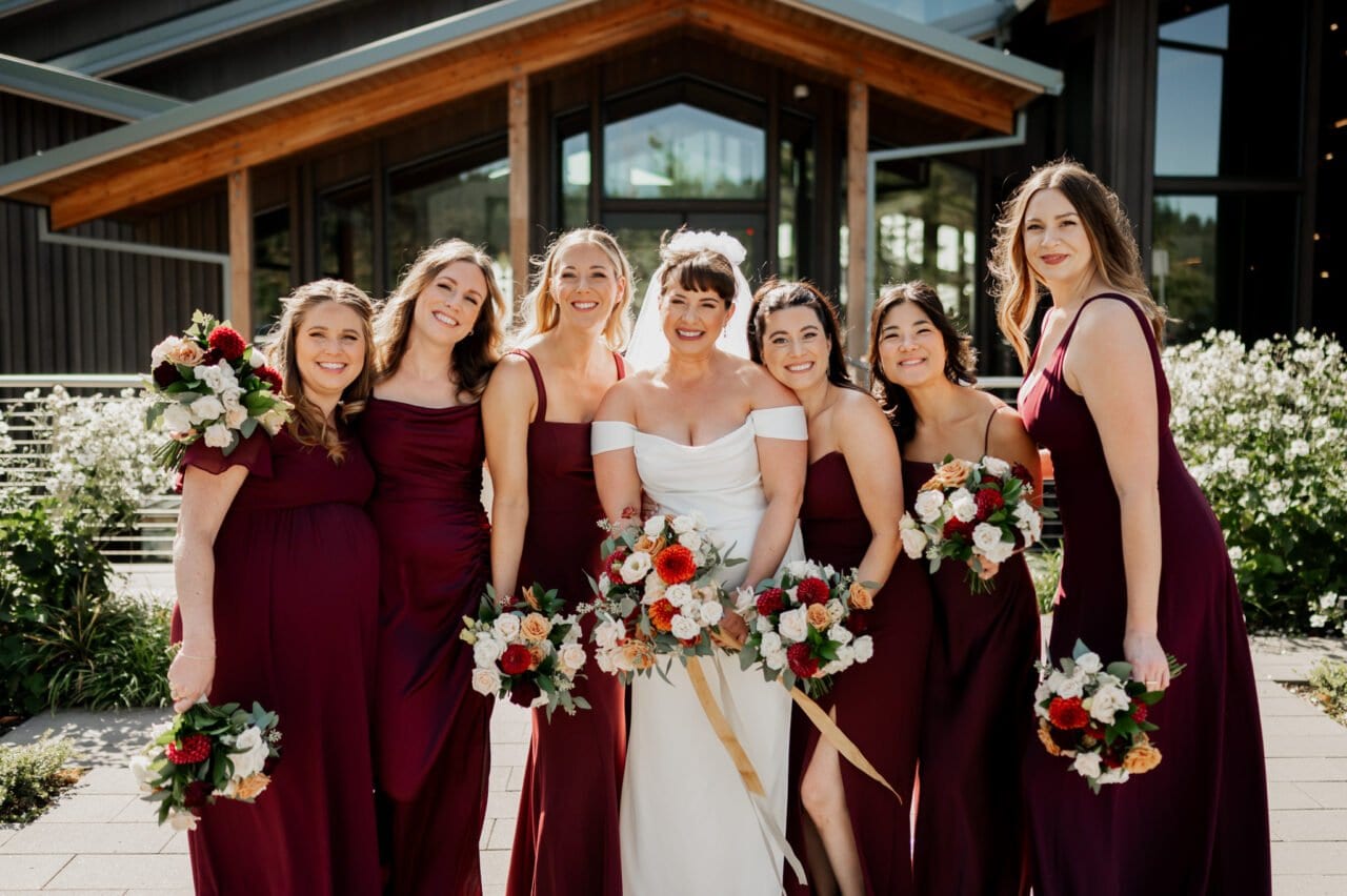 Bride in a stunning white dress with six bridesmaids in elegant burgundy gowns, each holding a bouquet, posed gracefully in front of Amaterra Winerys charming facade.