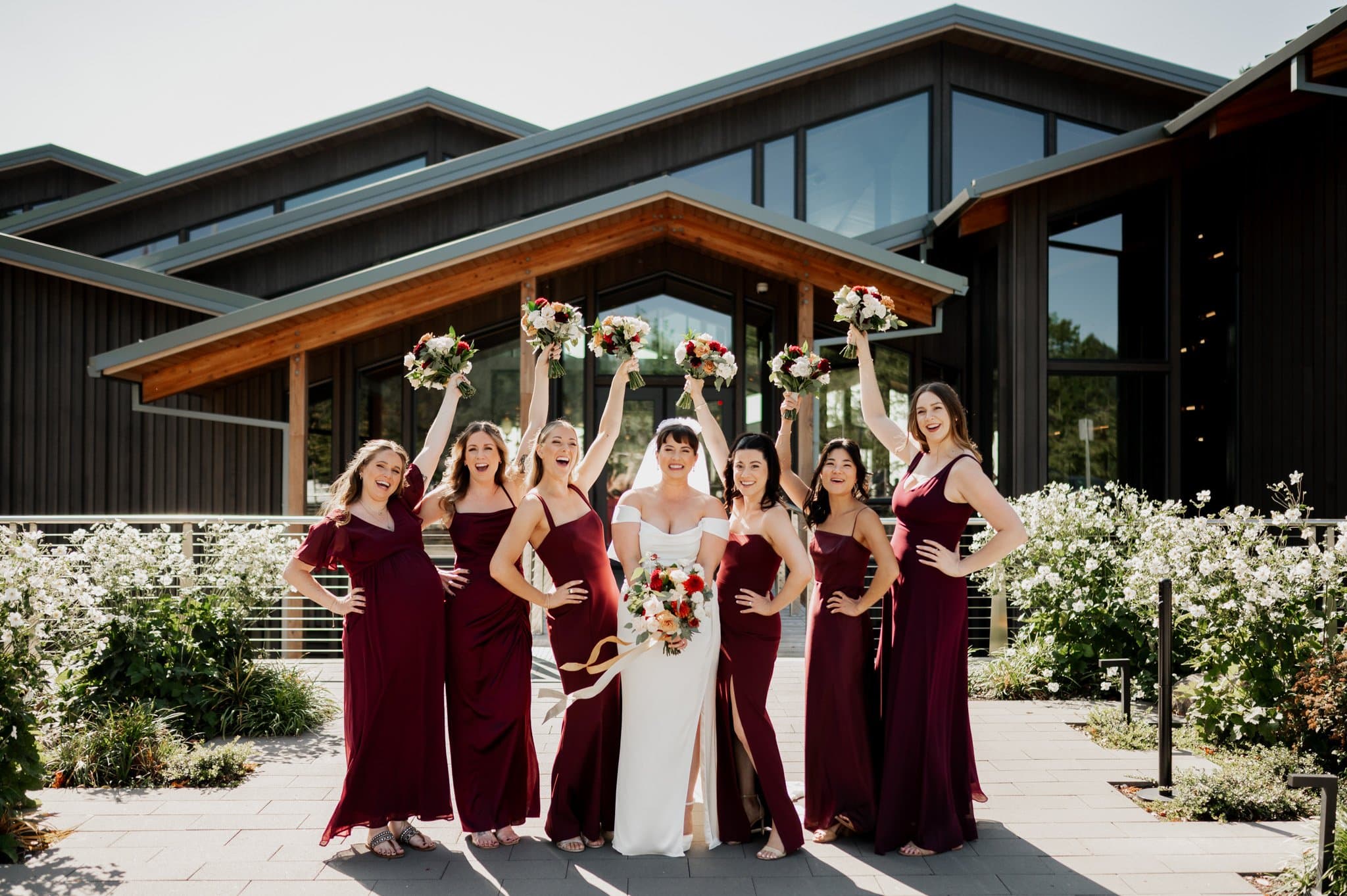 Bridal party posing joyfully at Amaterra Winery in burgundy dresses and a white gown, holding bouquets in front of a modern building.