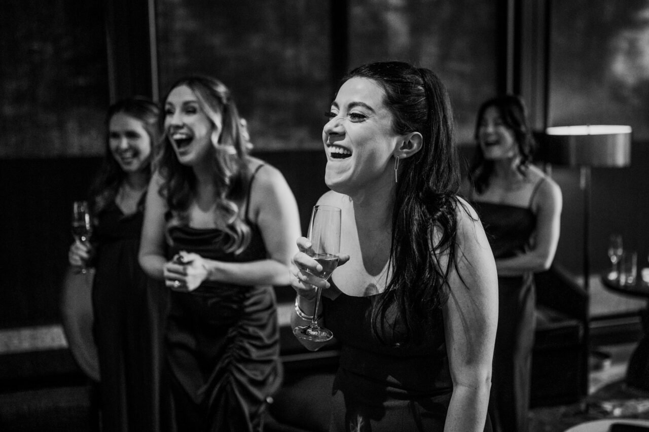 Four women in evening dresses laugh and hold glasses, savoring the vibrant atmosphere that feels like an evening at Amaterra Winery.