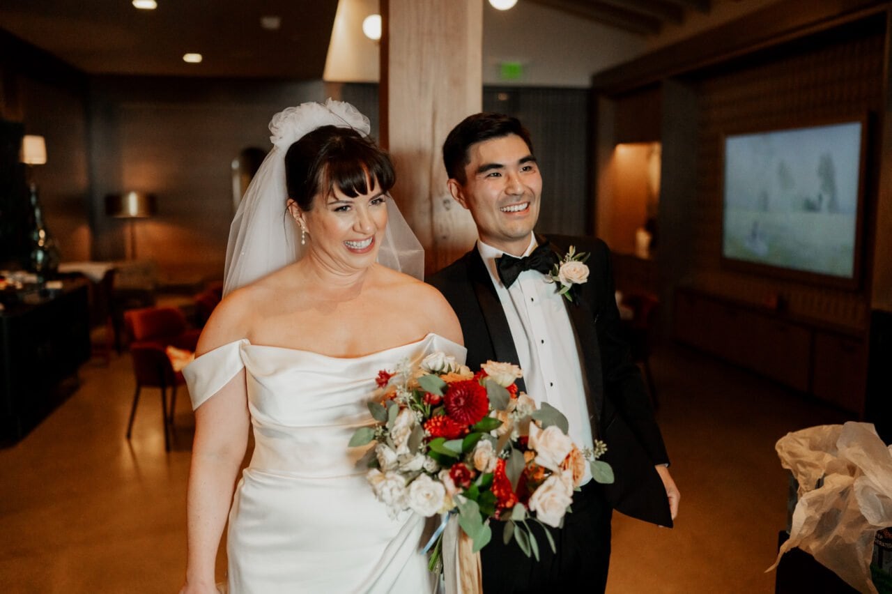 Bride in an off-shoulder gown and groom in a tuxedo smile, holding a bouquet with red and white flowers indoors, capturing the joy of their special day. This moment is beautifully framed at Amaterra Winery wedding photos.