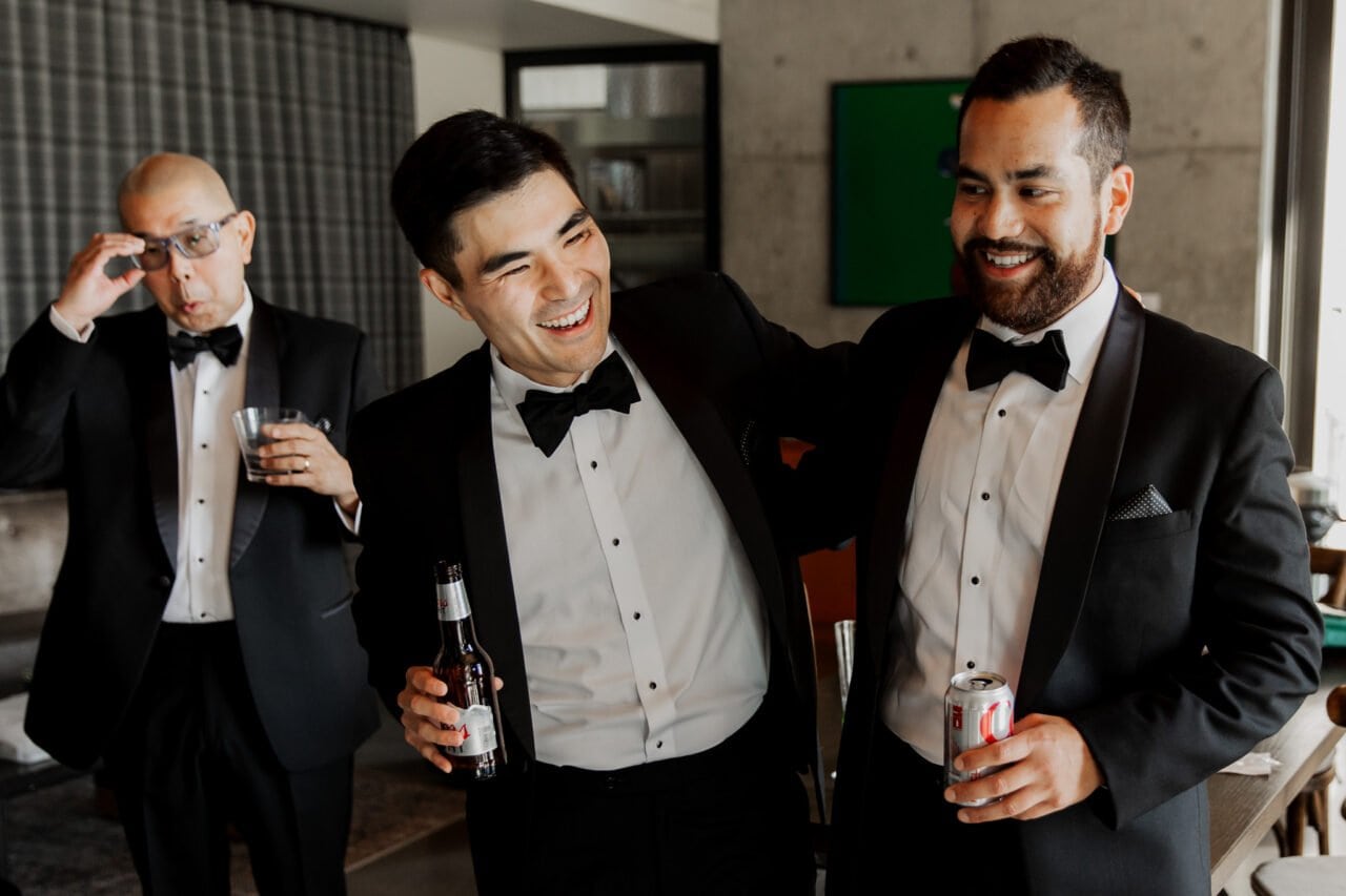 In a modern room, three men in tuxedos exude charm—two smiling and holding drinks as if captured straight from Amaterra Winery wedding photos, while the third adjusts his glasses in the background.