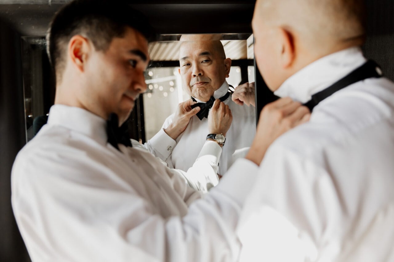 Two men in dress shirts fine-tune their bow ties in a mirror, intently focused on their reflections, as they prepare for the Amaterra Winery wedding photos.