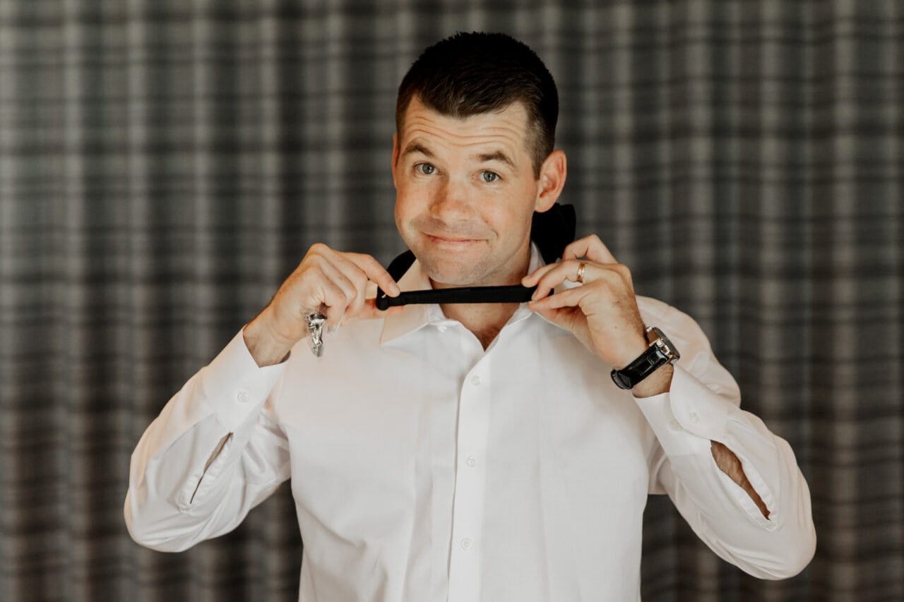 Man in white shirt adjusting bow tie, smiling slightly against a curtain backdrop—a picture perfect moment reminiscent of Amaterra Winery wedding photos.