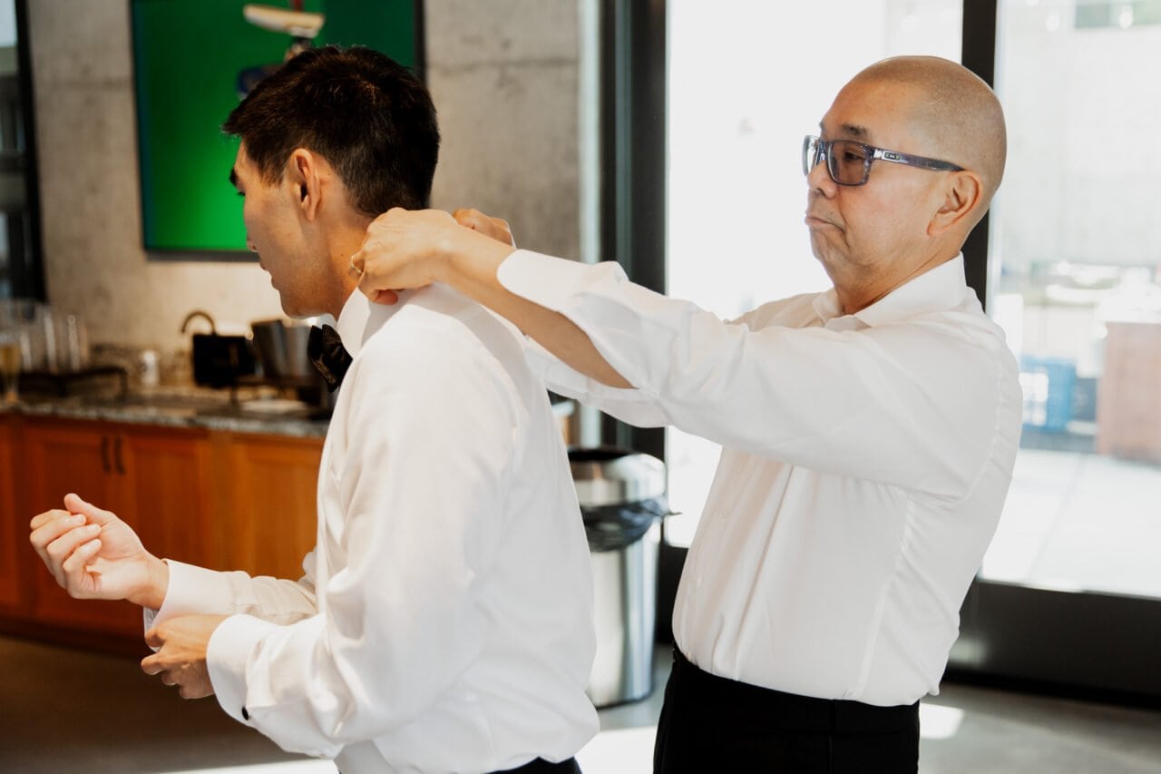 A man helps another adjust his tie in a bright room, the setting echoing the elegance of Amaterra Winery wedding photos. Both are dressed in crisp white shirts, capturing a moment of camaraderie and style.