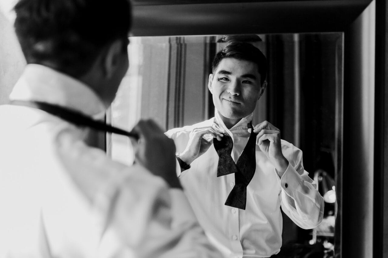 Man smiling while adjusting a bow tie in front of a mirror, wearing a white dress shirt, preparing for Amaterra Winery wedding photos.