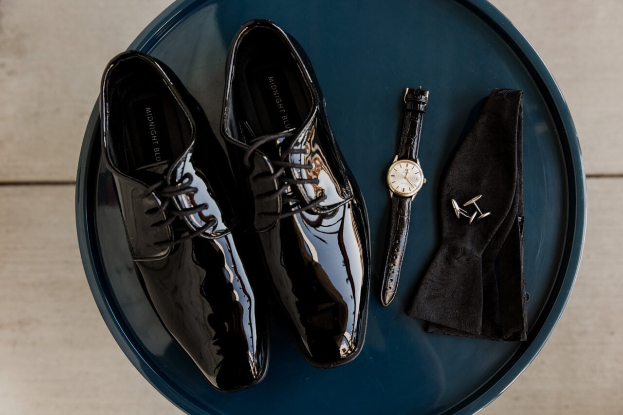 Shiny black dress shoes, a wristwatch, and cufflinks catch the light alongside a folded pocket square on a round blue table, setting the scene for elegant Amaterra Winery wedding photos.