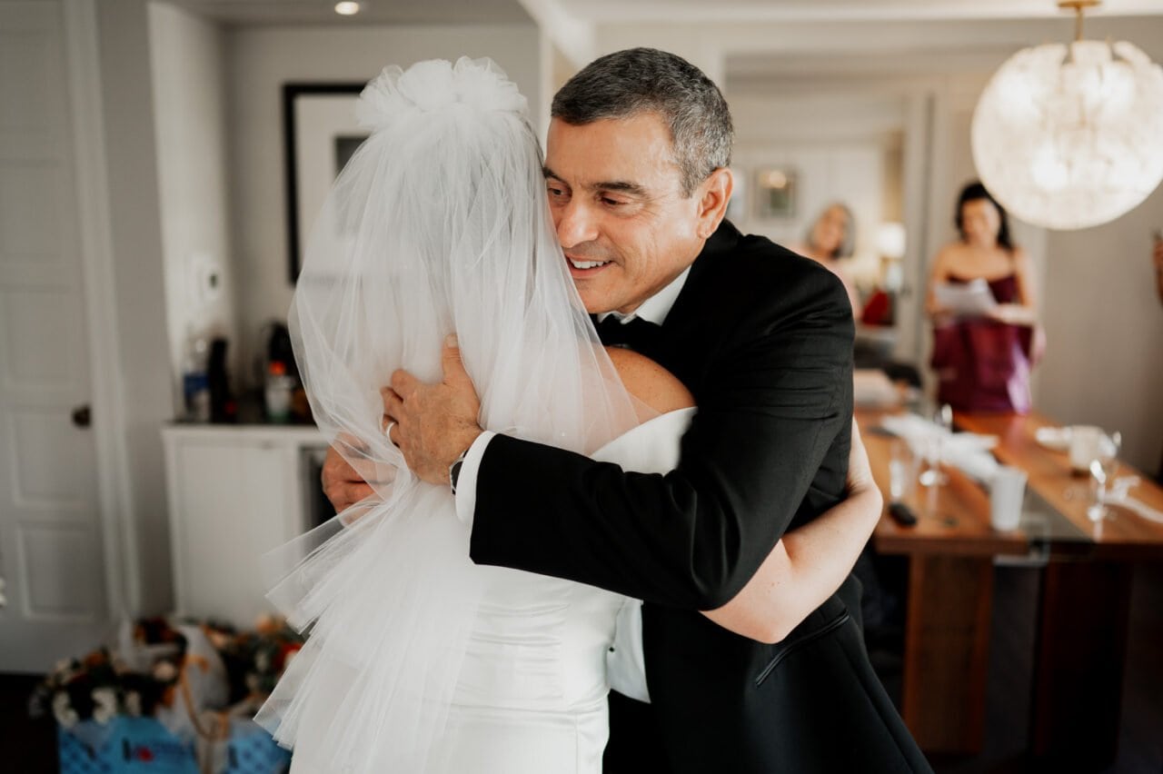 A bride in a white dress and veil hugs a man in a suit in a warmly lit room, getting ready at The Heathman Hotel for an Amaterra Winery wedding.