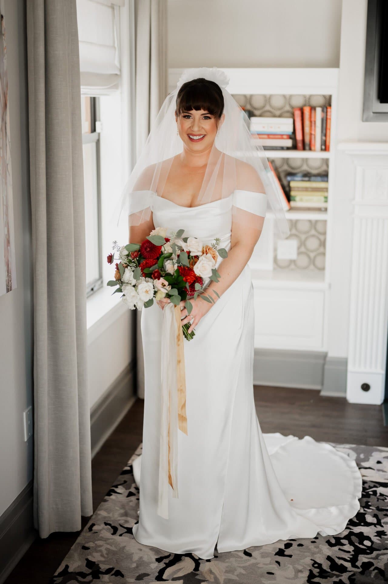 The bride, getting ready at The Heathman Hotel, stands gracefully indoors near a window, her white dress elegantly draped as she clutches a bouquet. Behind her, bookshelves add an air of sophistication, setting the perfect backdrop before heading to Amaterra Winery for the wedding.