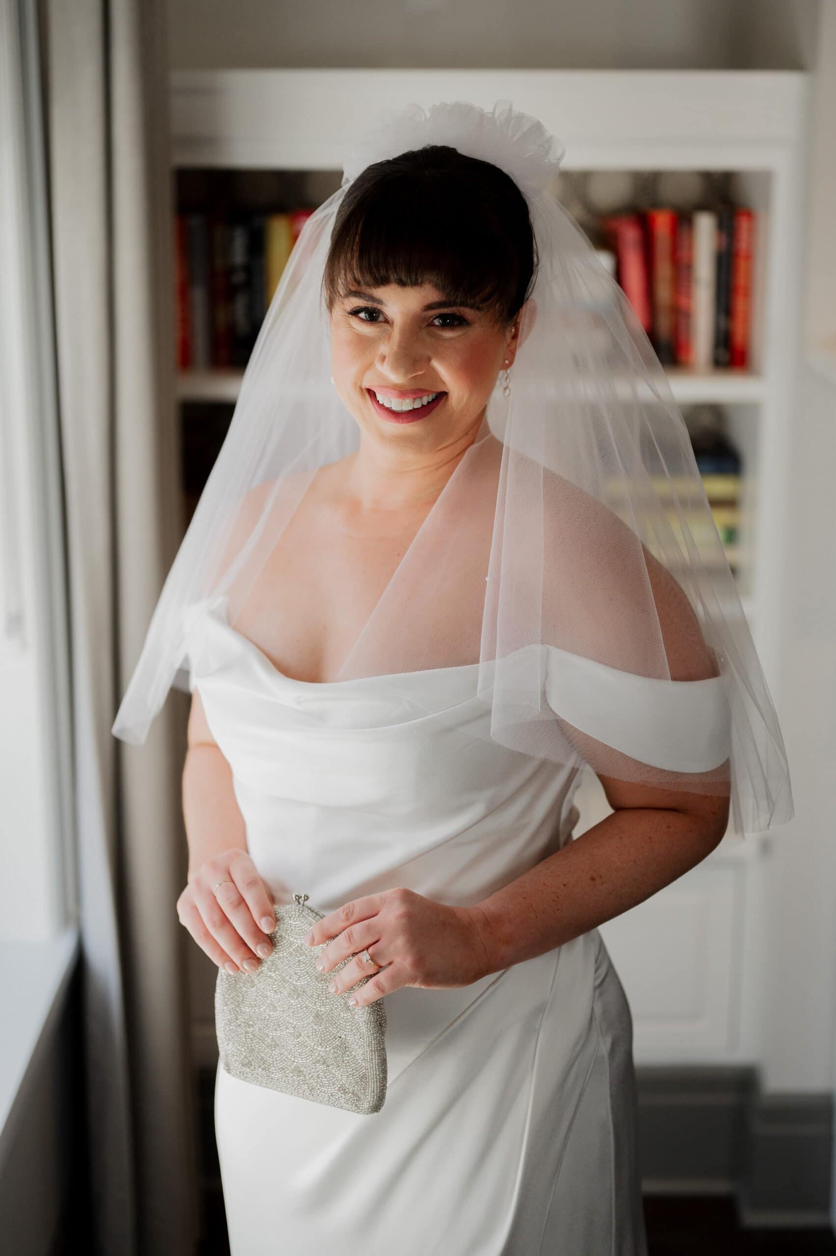 The bride, in an elegant white dress and veil, holds a small clutch and smiles while getting ready at The Heathman Hotel. A bookshelf stands in the background, hinting at her upcoming Amaterra Winery wedding.