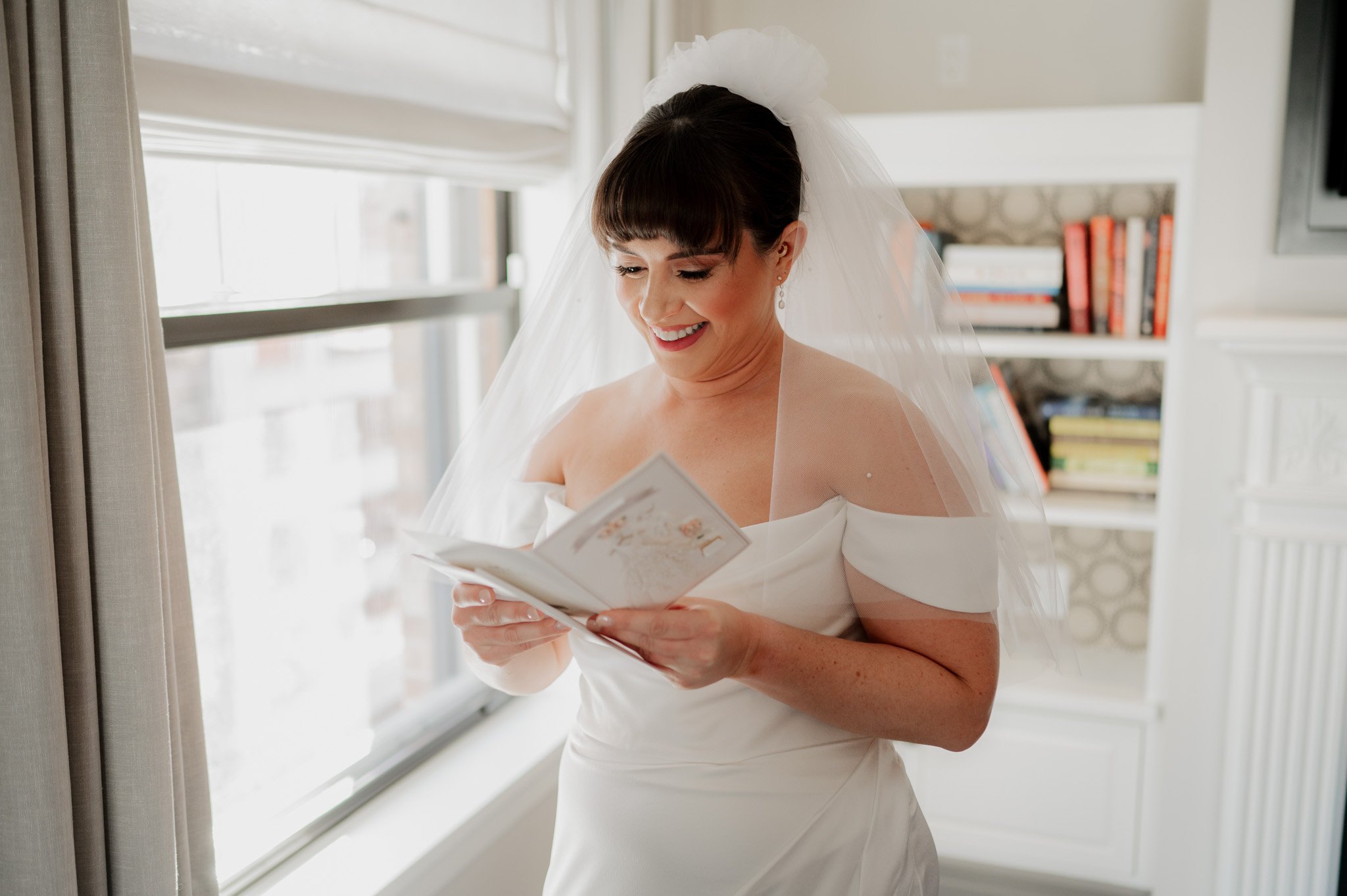 The bride, draped in a white dress and veil, reads a card by the window of The Heathman Hotel as she gets ready for her Amaterra Winery wedding. A bookshelf stands gracefully in the background.