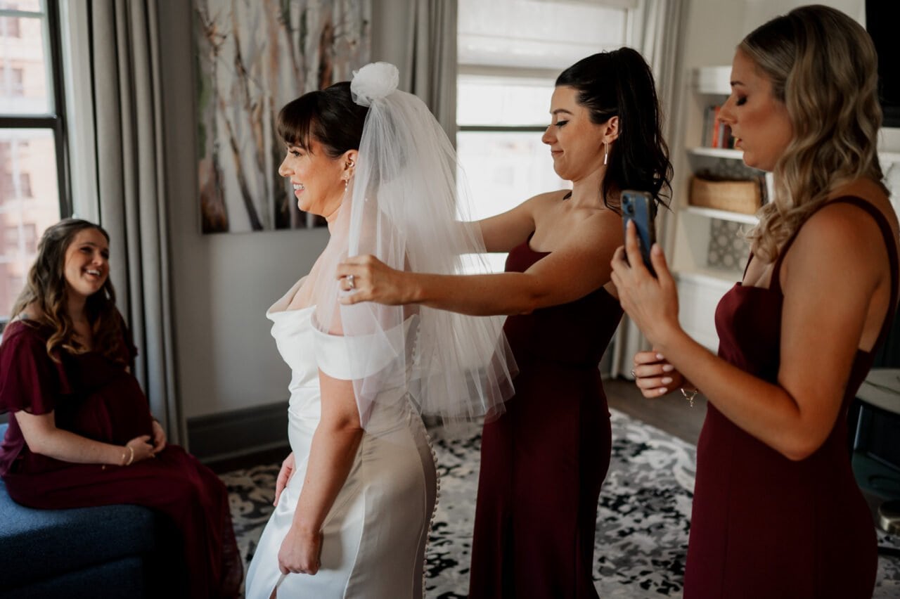 The bride gets ready with her bridesmaids at The Heathman Hotel, adjusting her veil and snapping pictures, as another woman watches from the couch.