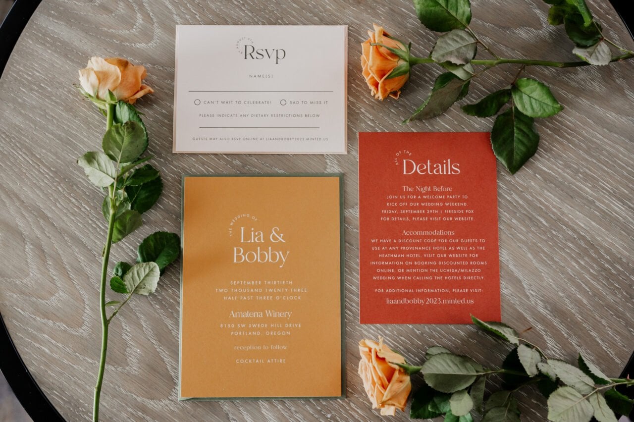 Wedding invitations and RSVP cards rest on a textured surface, surrounded by vibrant orange roses, setting the tone for a beautiful day getting ready at The Heathman Hotel for an elegant Amaterra Winery wedding.