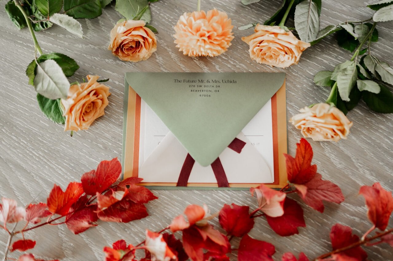 A green envelope surrounded by orange roses and red leaves rests elegantly on a wooden surface, capturing the anticipation of getting ready at The Heathman Hotel for an Amaterra Winery wedding.