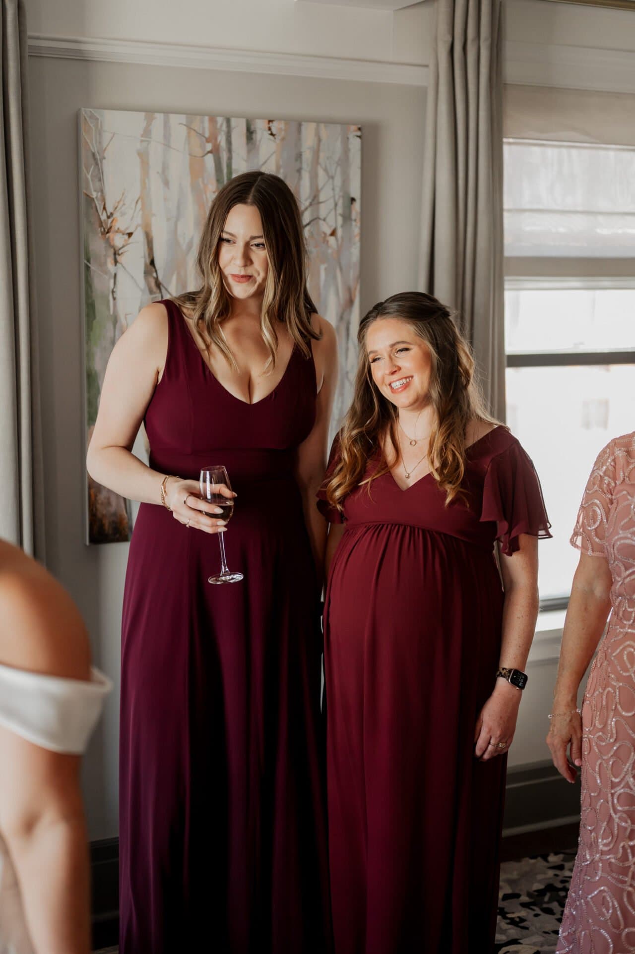 Two women in matching burgundy dresses smile indoors as they get ready at The Heathman Hotel for an Amaterra Winery wedding. One holds a wine glass, with a painting in the background adding to the elegant ambiance.